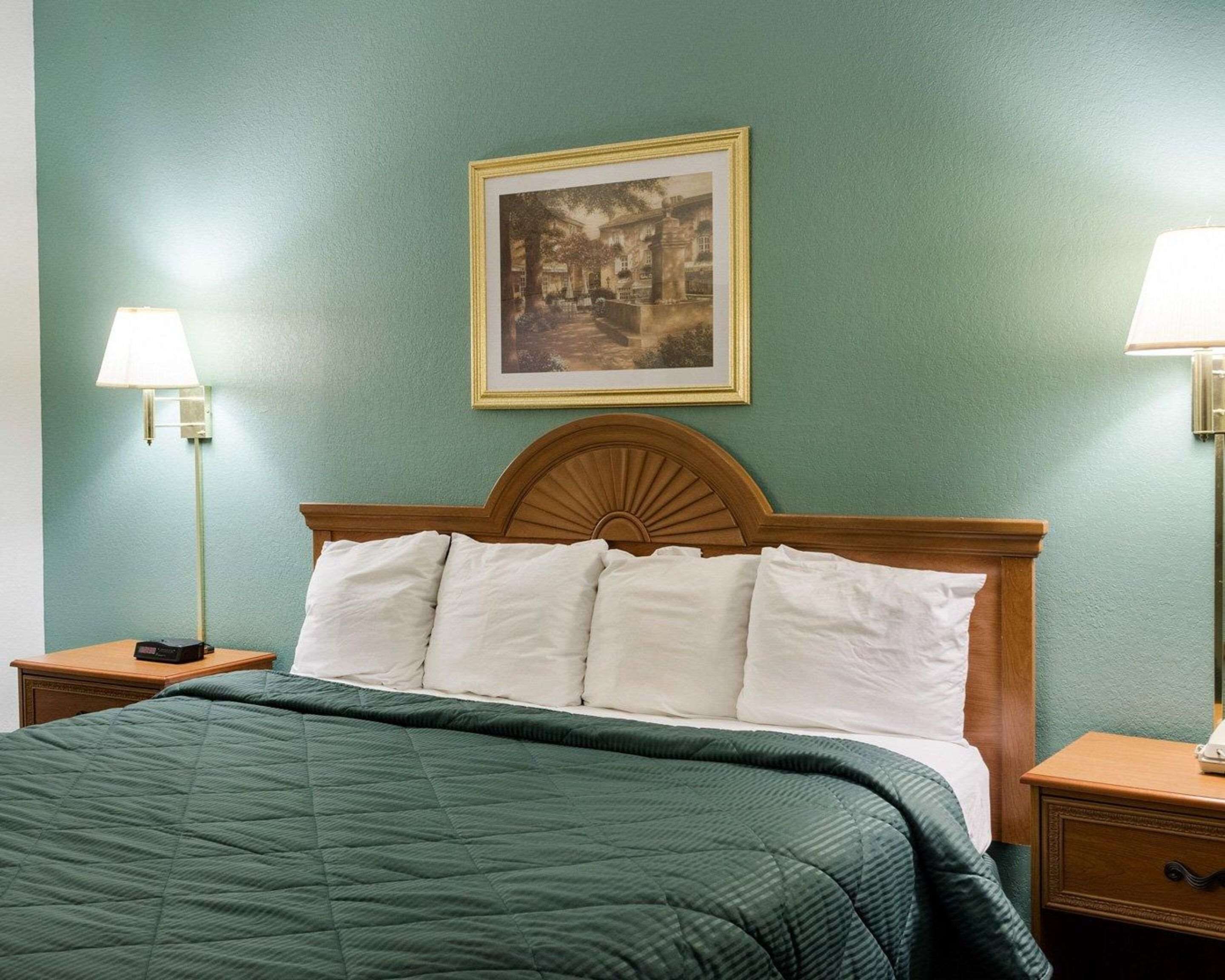 A neatly made bed with a green quilt, two bedside lamps, a framed picture on the wall, and a wooden headboard.