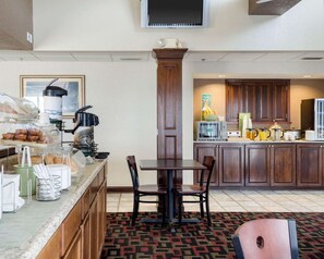 Et område til hotellets morgenmad med buffet, kaffemaskine og mikroovn. Et spisebord med stole og et mønstret tæppe.