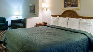 A hotel room with a large bed, a nightstand with a lamp, a chair, a framed picture on the wall, and a carpeted floor. - Quality Inn Magee (Magee)