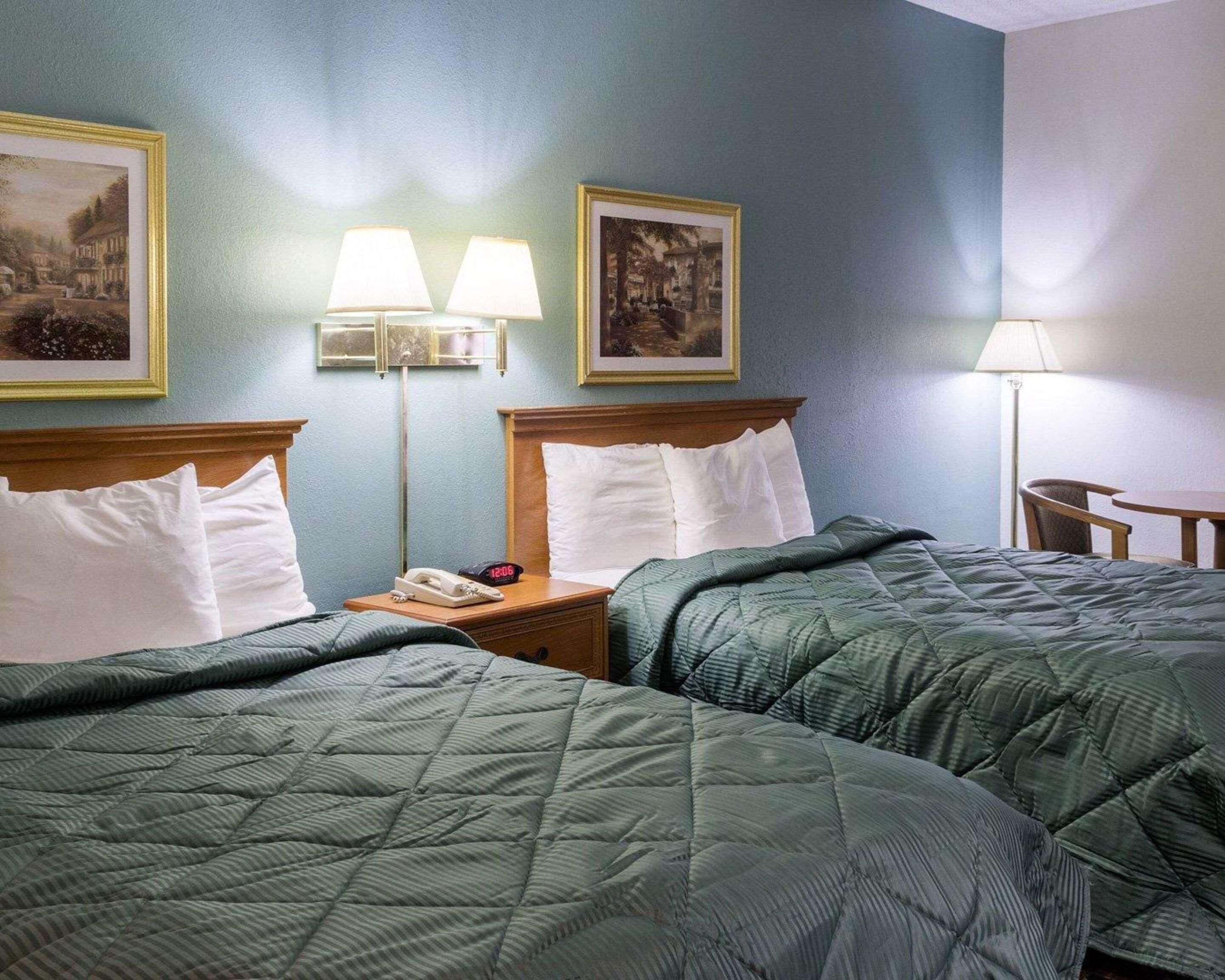 A hotel room with two beds, a nightstand with a phone and clock, and two wall-mounted lamps.