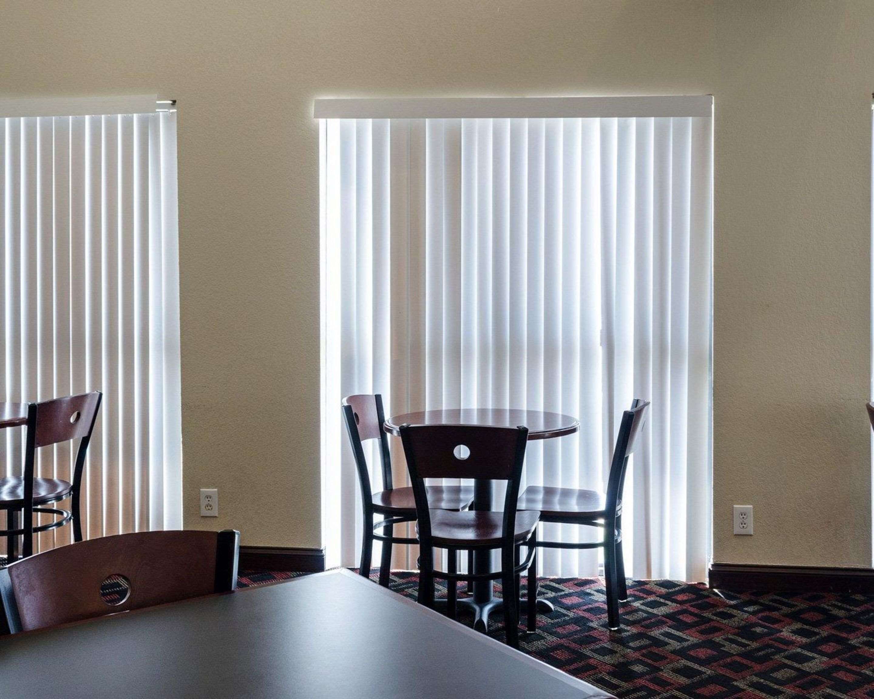 A hotel room with a dining area featuring a table and four chairs, a patterned carpet, and a large window with vertical blinds.