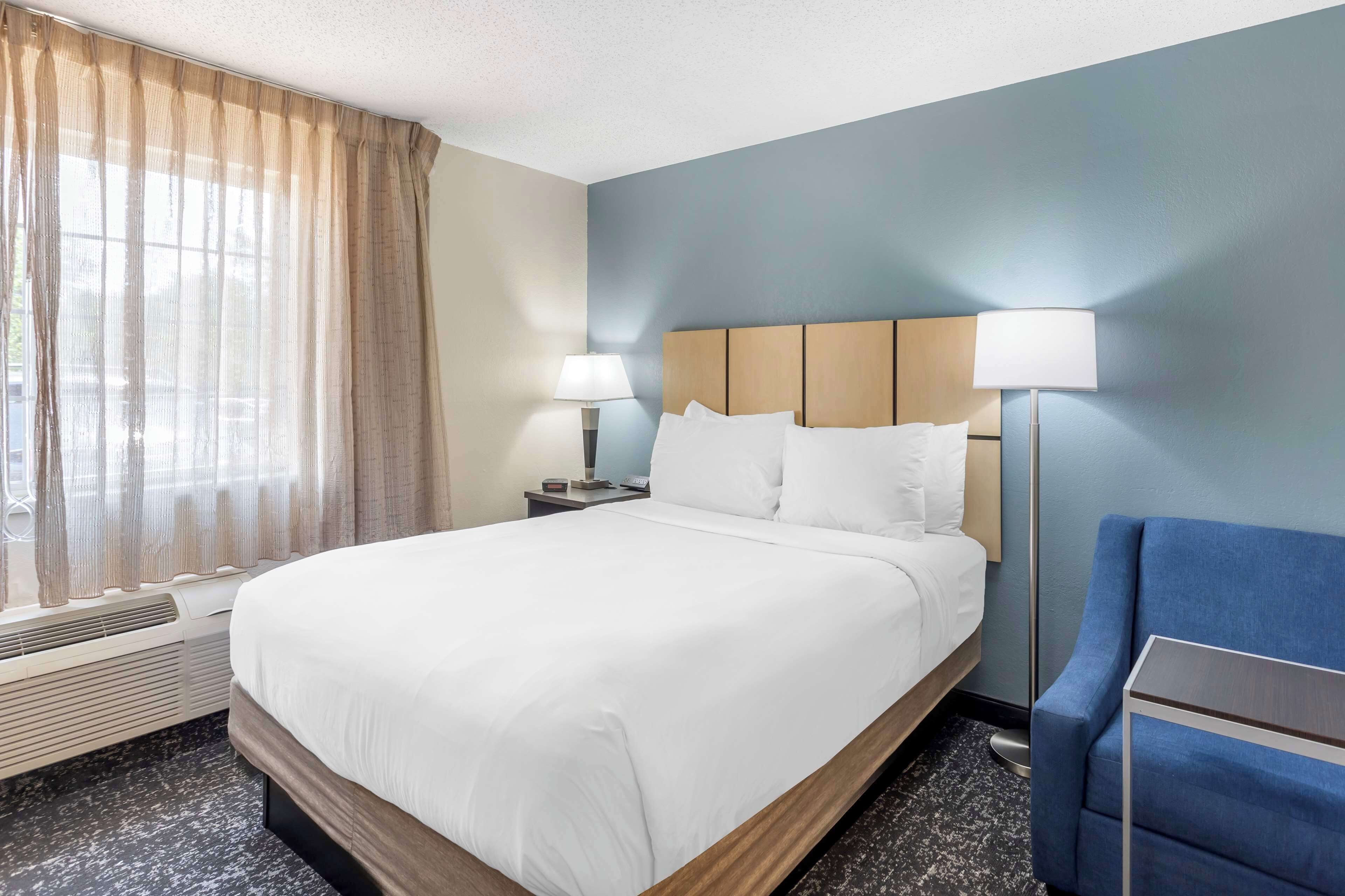 A hotel room with a large bed, a desk with a lamp, a chair, and a window with curtains.