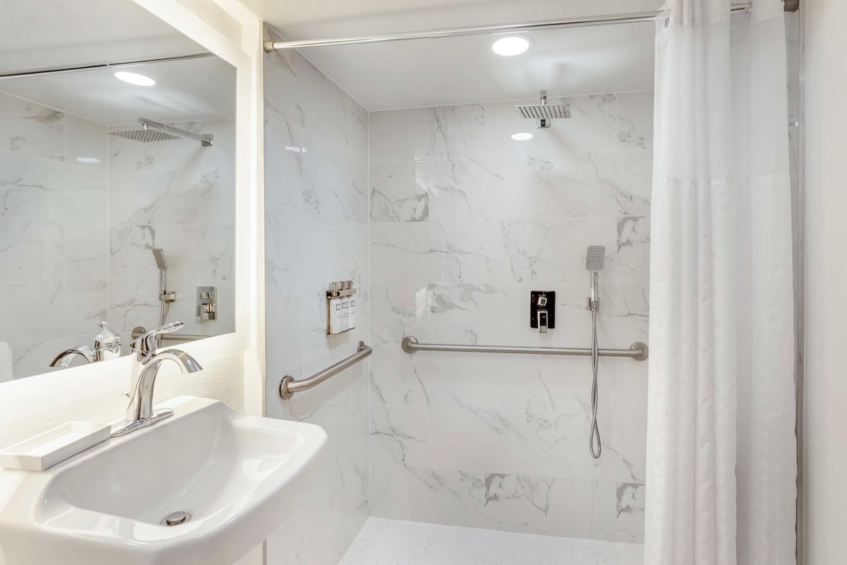 A modern bathroom with a white sink, marble walls, a shower with a handheld showerhead, and a handrail.