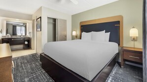 Habitación de hotel con una cama grande, una mesita de noche, un tocador, un ventilador de techo, un baño con espejo y una puerta.