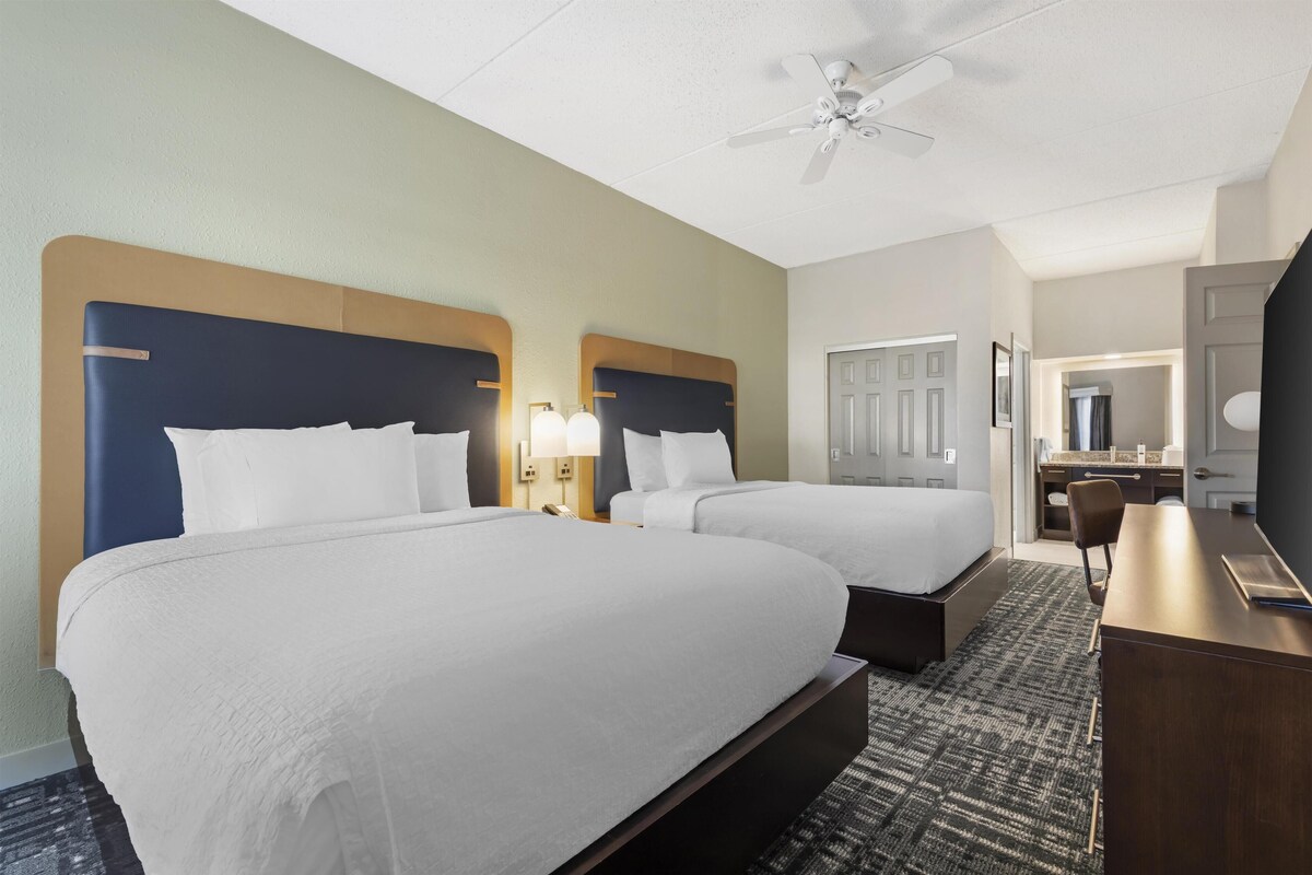 A hotel room with two beds, a desk, a chair, a television, and a ceiling fan.