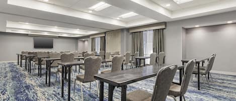 A conference room with multiple tables and chairs, a flat-screen TV on the wall, and a patterned carpet.