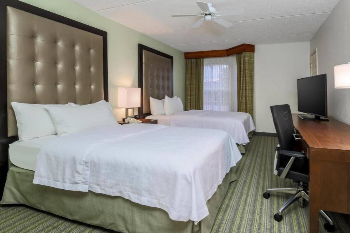 A hotel room with two beds, a desk, a chair, a television, and a ceiling fan.