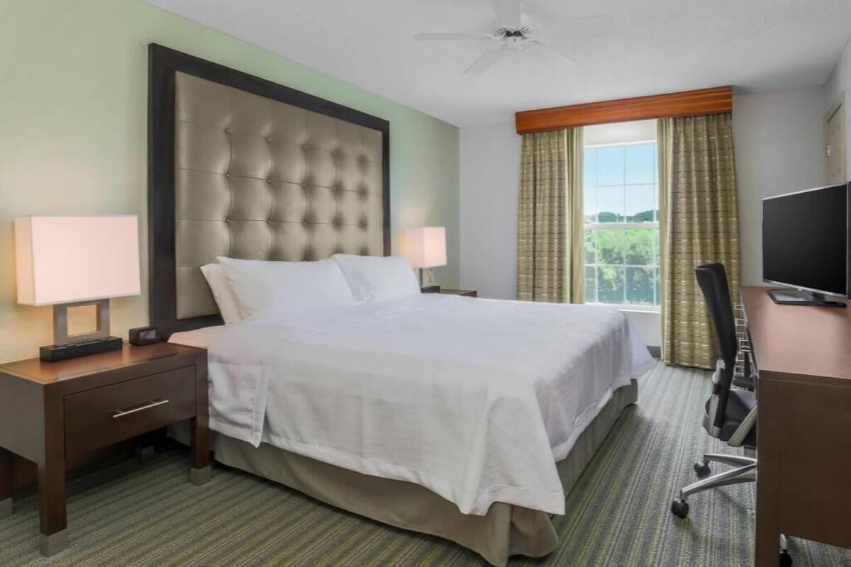 A hotel room with a large bed, a desk with a computer, a chair, a TV, and a window with curtains.