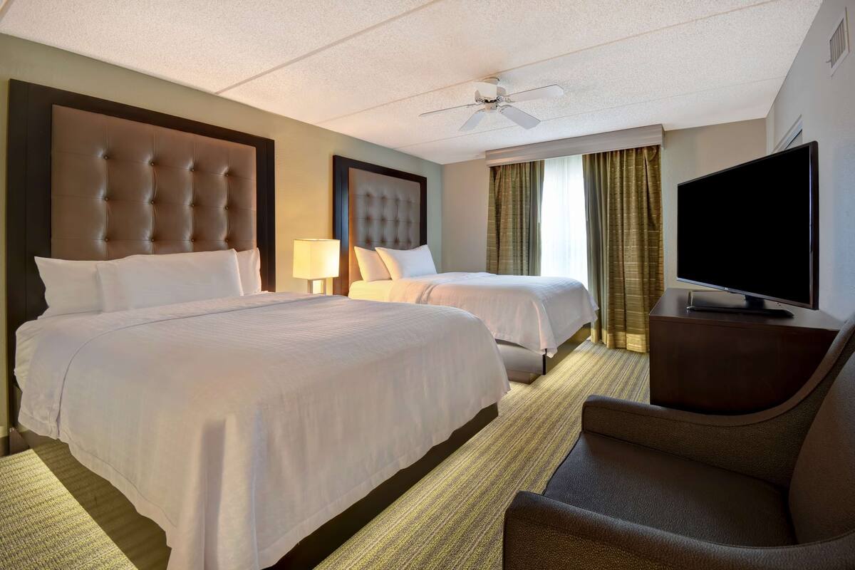 A hotel room with two beds, a television, a ceiling fan, and a large window with curtains.
