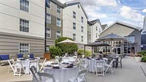 An outdoor patio area with round tables and chairs, some with umbrellas, set against a building with multiple windows and a flat roof. - Homewood Suites by Hilton Philadelphia Great Valley (Malvern)