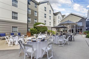 An outdoor patio area with round tables and chairs, some with umbrellas, set against a building with multiple windows and a flat roof. - Homewood Suites by Hilton Philadelphia Great Valley (Malvern)