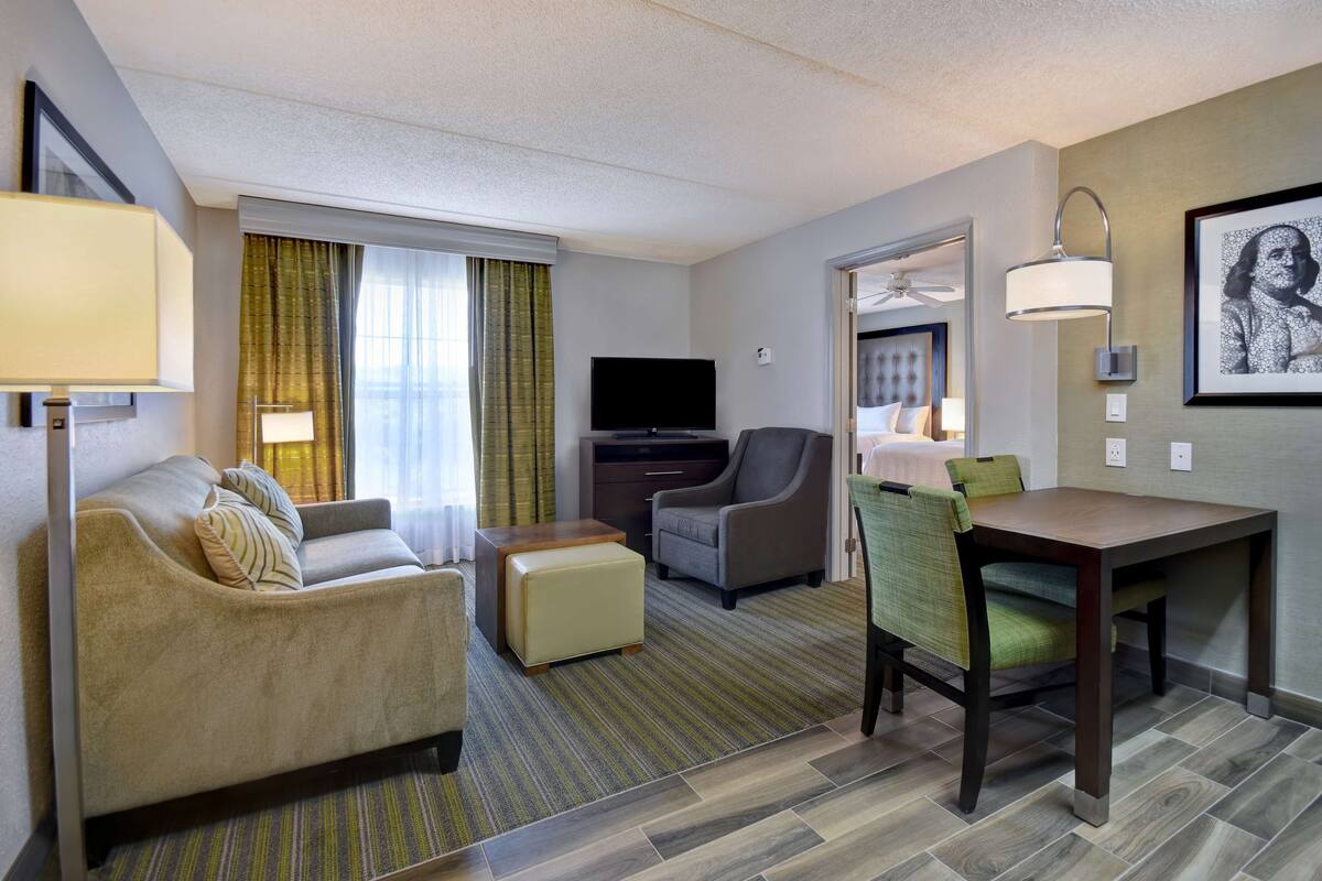 A hotel room with a sofa, armchair, ottoman, TV, and a dining table with chairs.