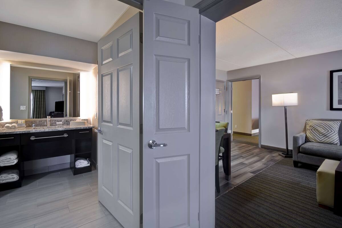 A hotel room with a bathroom, a sitting area with a sofa and a lamp, and a hallway leading to another room.