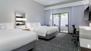 Ein Hotelzimmer mit zwei Betten, einem Schreibtisch, einem Stuhl, einem Fernseher und einer Schiebeglasdoor zur Außenwelt.