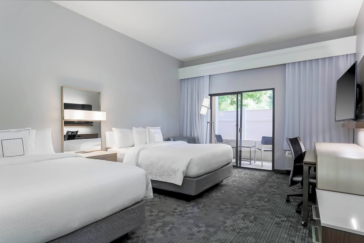 A hotel room with two beds, a desk, a chair, a TV, and a sliding glass door to the outside.