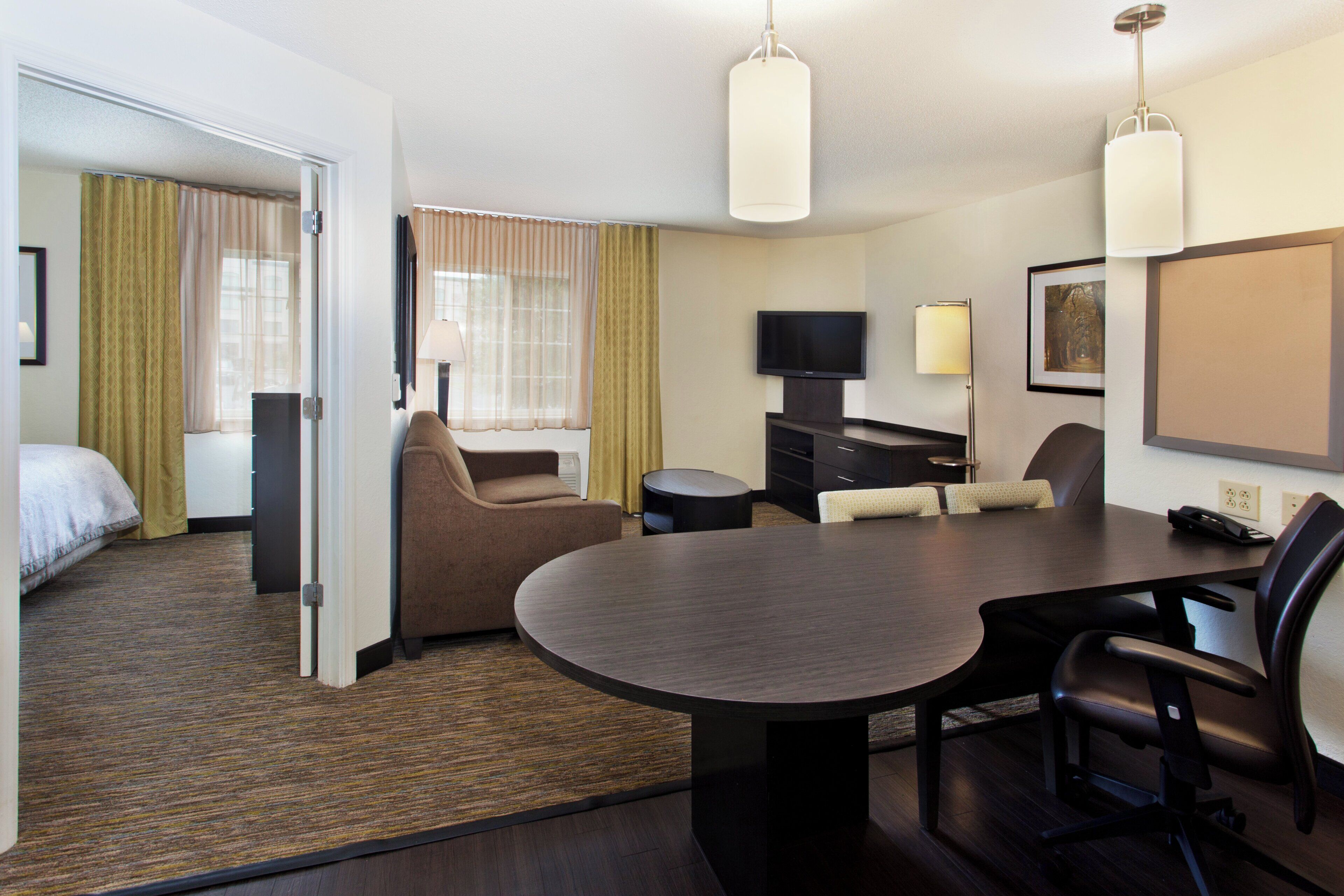 Habitación de hotel con cama, zona de estar con mesa y sillas, televisor y escritorio con lámpara.