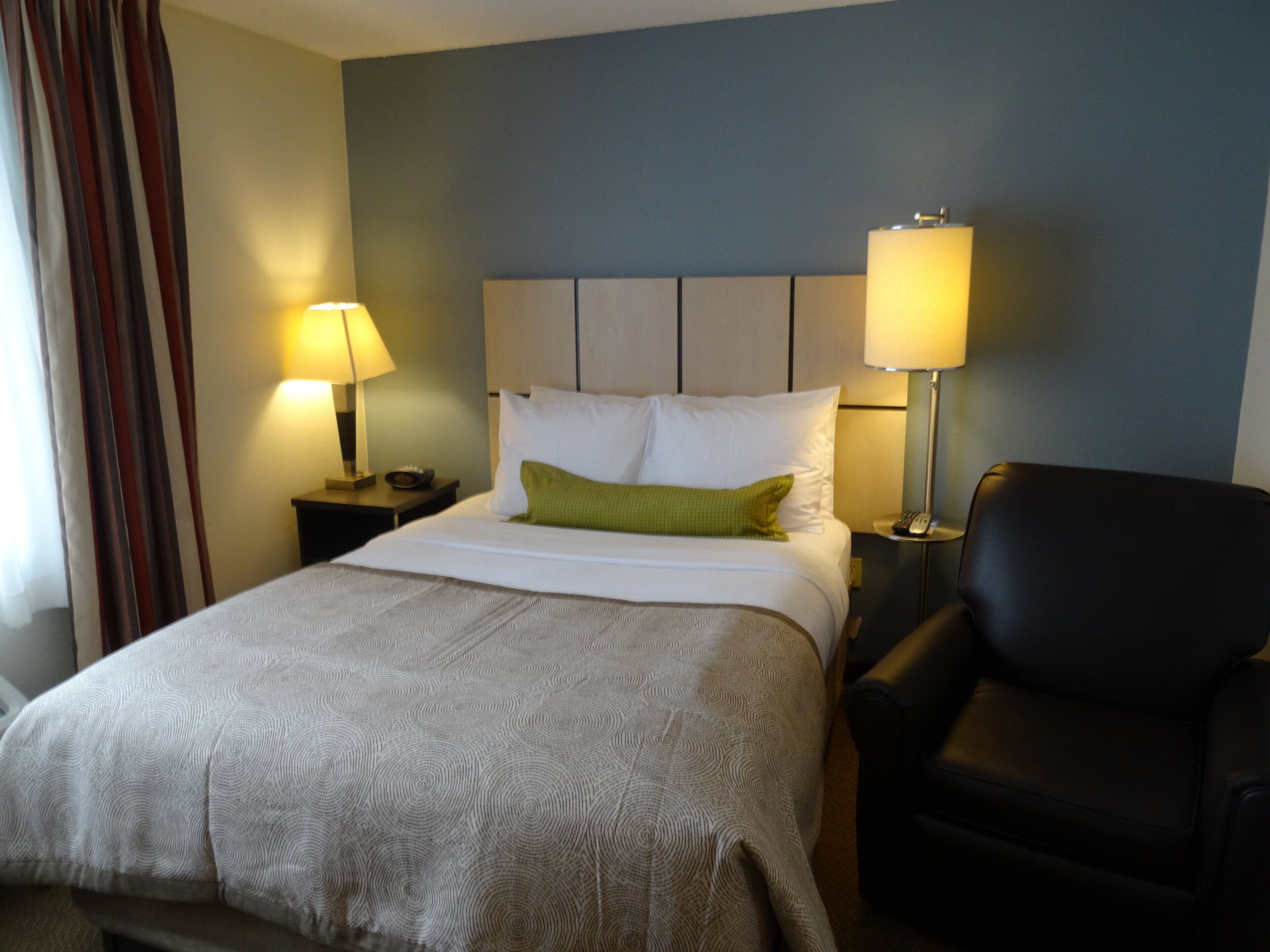A hotel room with a bed, two bedside lamps, a green pillow, a black chair, and a nightstand with a remote control.