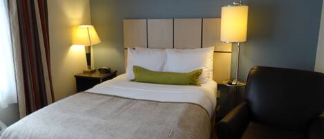 Habitación de hotel con cama, dos lámparas de noche, almohada verde, silla negra y mesita de noche con control remoto.
