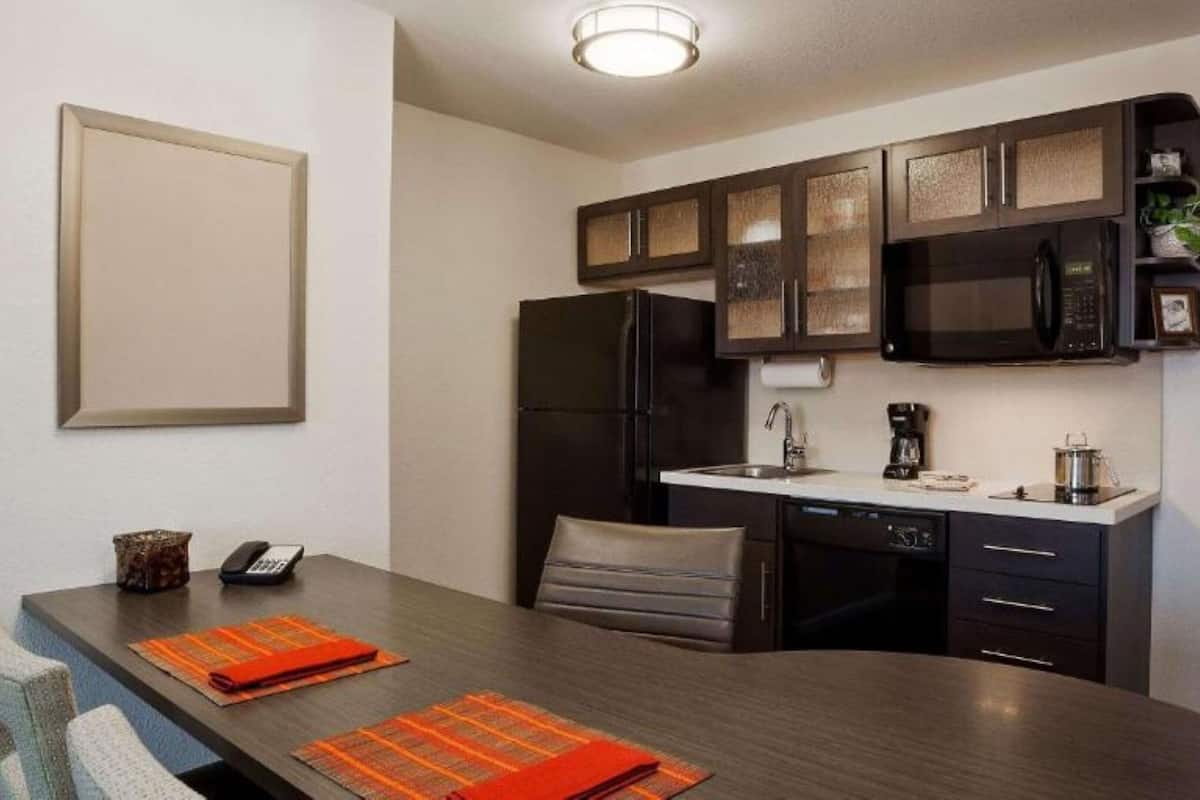 A compact kitchen with dark cabinets, a refrigerator, a microwave, and a coffee maker. There is a dining table with two chairs and orange placemats.