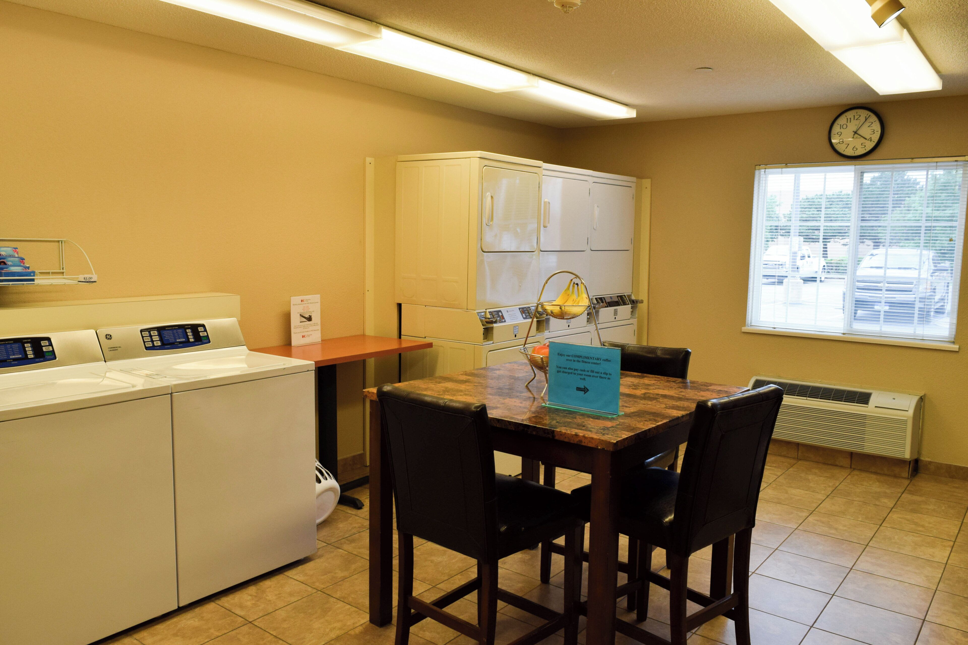 A hotel room with a kitchenette, a dining area with a table and chairs, a clock on the wall, and a window with blinds.
