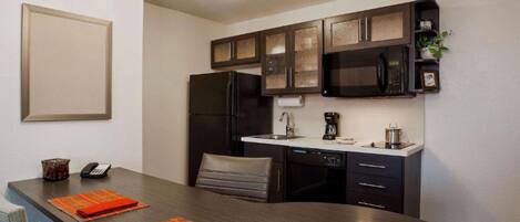 A compact kitchen with dark cabinets, a refrigerator, a microwave, and a coffee maker. There is a dining table with two chairs and orange placemats.