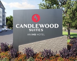 Letrero de Candlewood Suites, un hotel IHG, con logotipo, nombre y vista del área de estacionamiento y follaje.