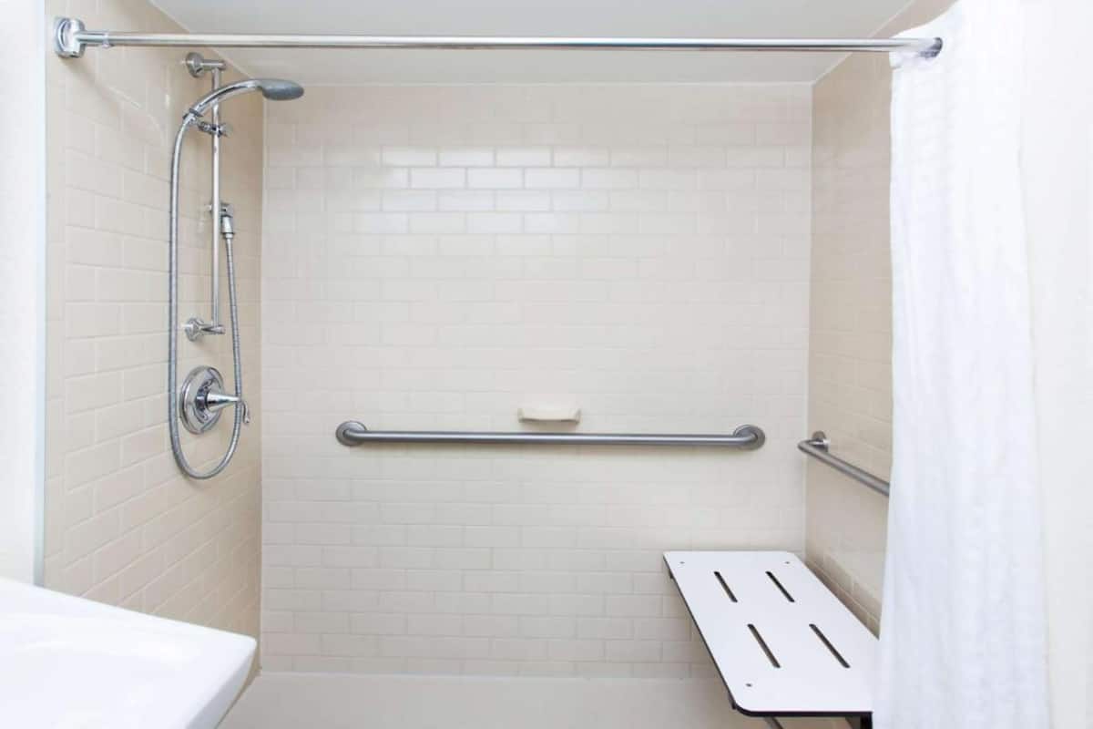 A shower with a grab bar, a white bathtub, and a white tiled wall.