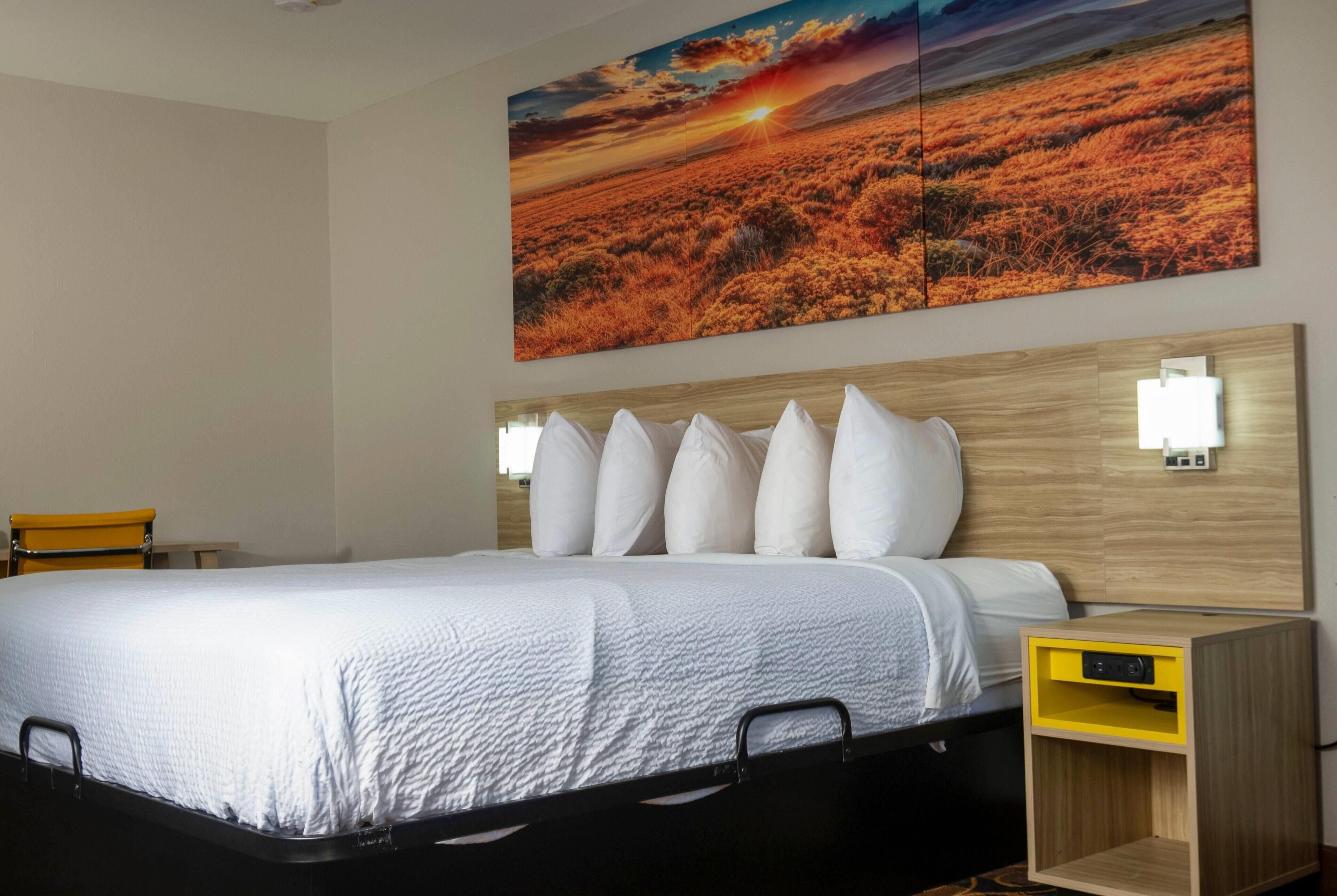A hotel room with a large bed, a headboard with lights, a nightstand, and a wall-mounted painting.