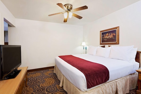 A hotel room with a bed, a television, a ceiling fan, a painting on the wall, and a patterned carpet.