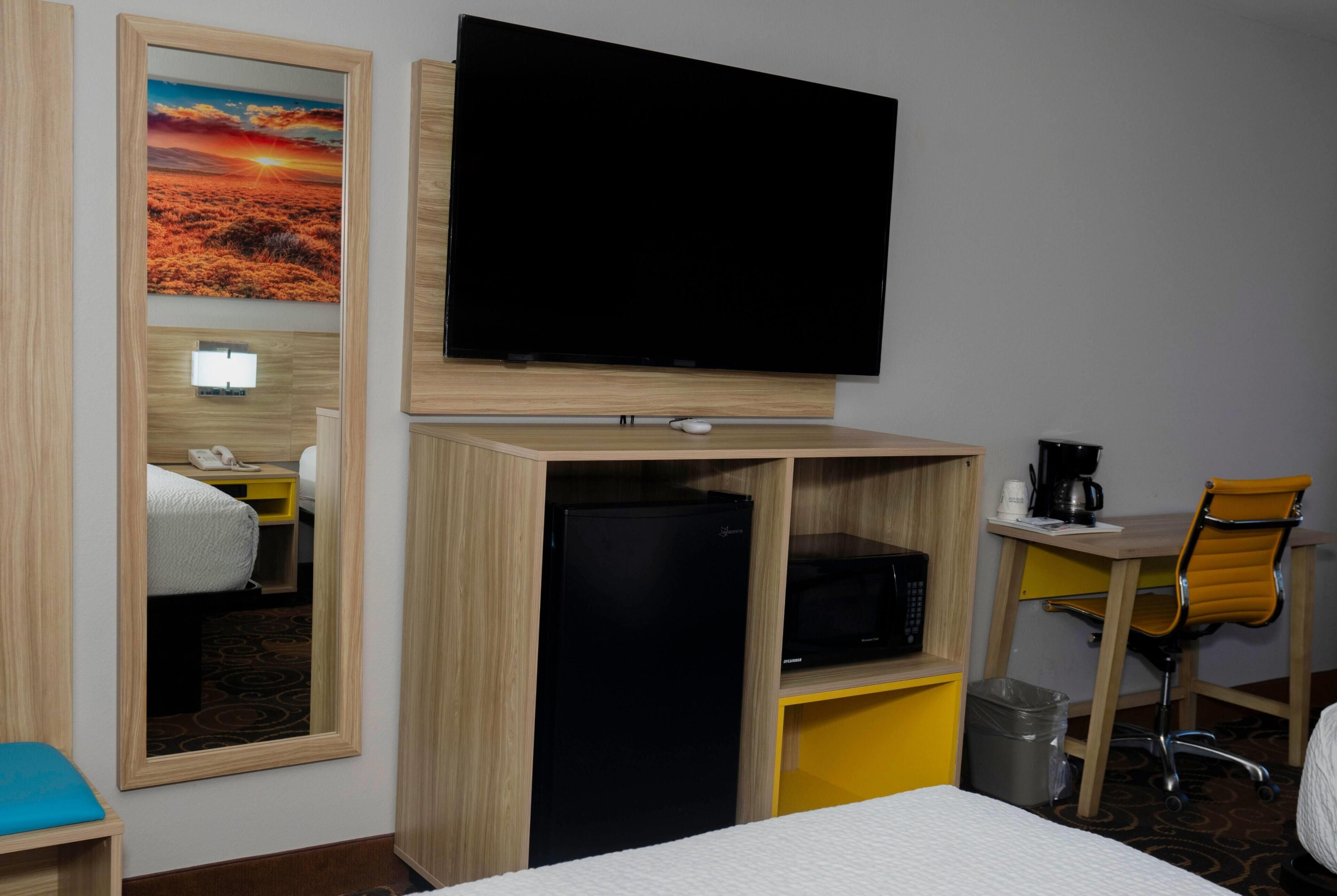 Una camera d'albergo con un letto, una scrivania con una sedia, una televisione, un frigorifero e un cestino.