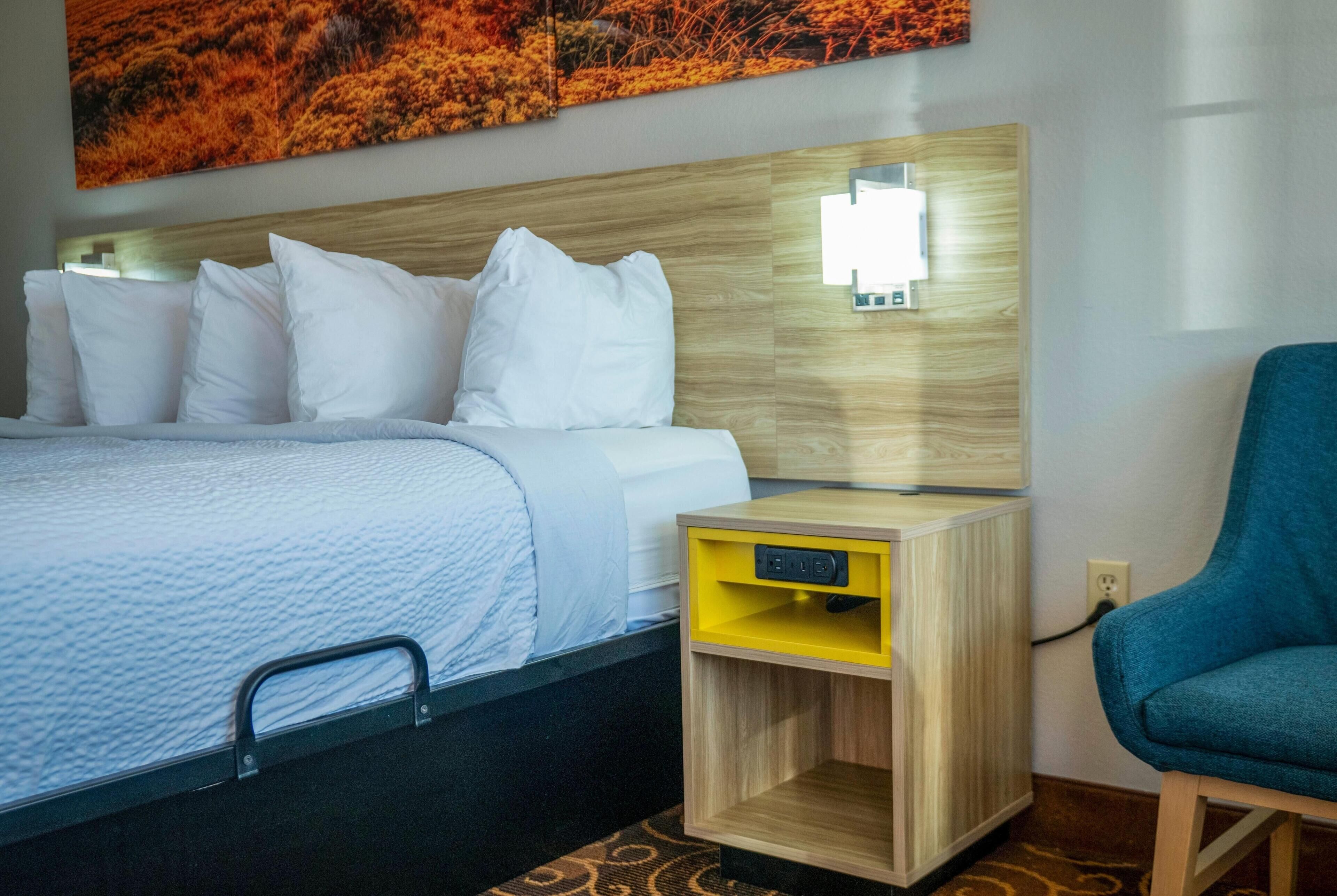 A hotel room with a bed, a bedside table with a lamp, a chair, and a wall-mounted artwork.