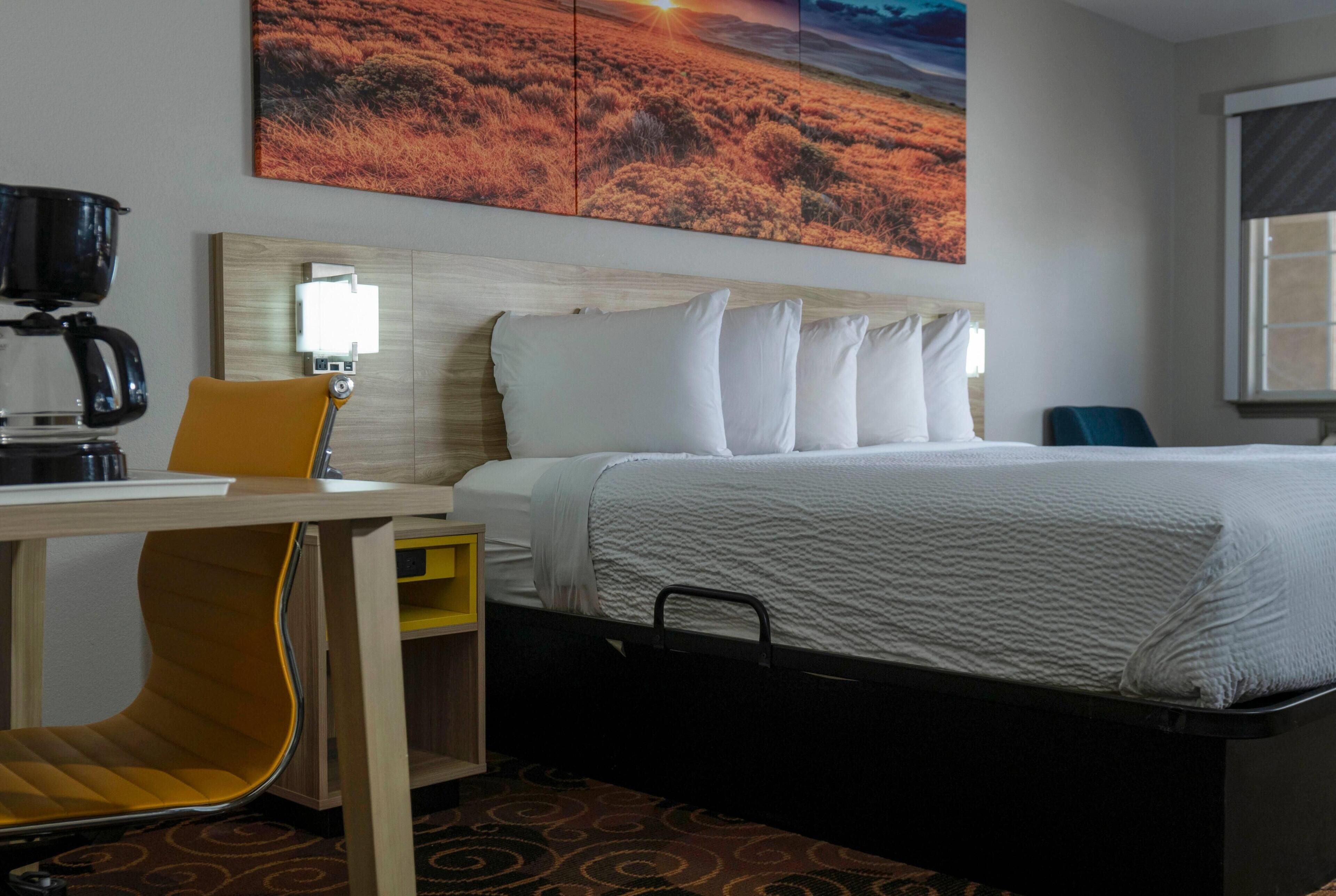 Habitación de hotel con cama, escritorio con cafetera, silla, lámpara y un cuadro en la pared.