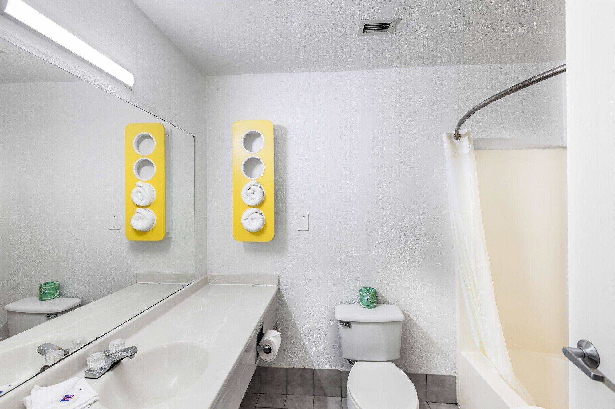 Une salle de bain moderne avec un lavabo blanc, un miroir et des toilettes. On y trouve deux porte-serviettes avec des supports jaunes et une douche avec un rideau.