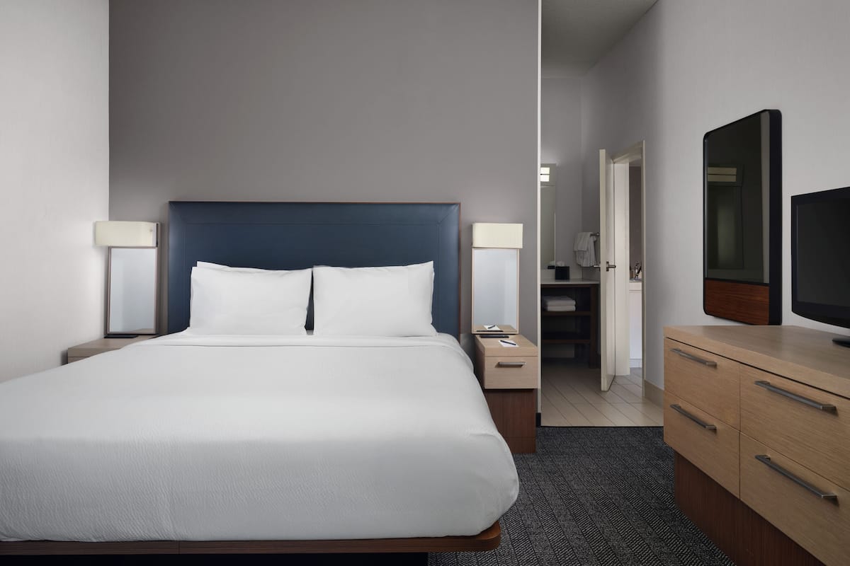 Een net opgemaakt bed met witte lakens, een houten nachtkastje met een lamp, een houten dressoir met een spiegel en een televisie.
