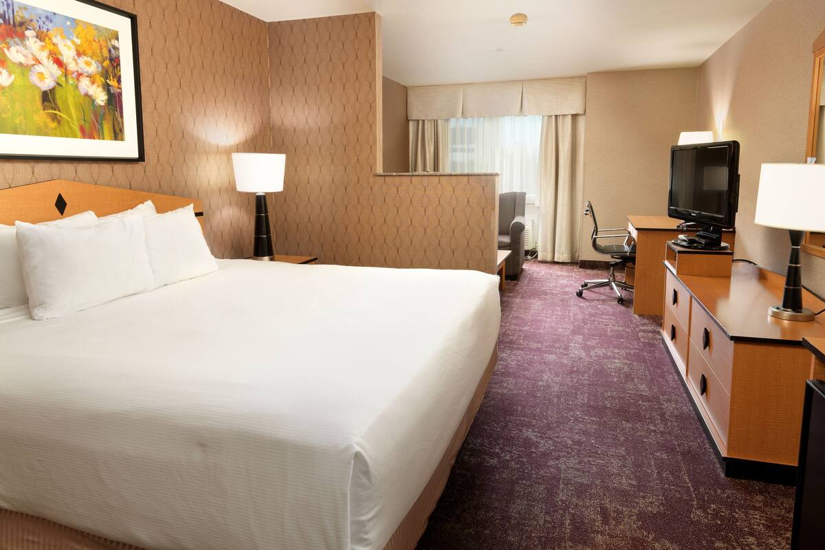 A hotel room with a large bed, a desk with a television, a chair, and a desk lamp.