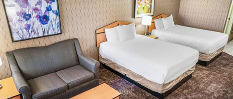 A hotel room with a sofa, two beds, a small table, and artwork on the walls.
