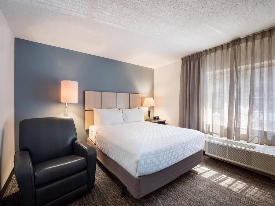 A hotel room with a bed, a chair, a nightstand with lamps, and a window with curtains.