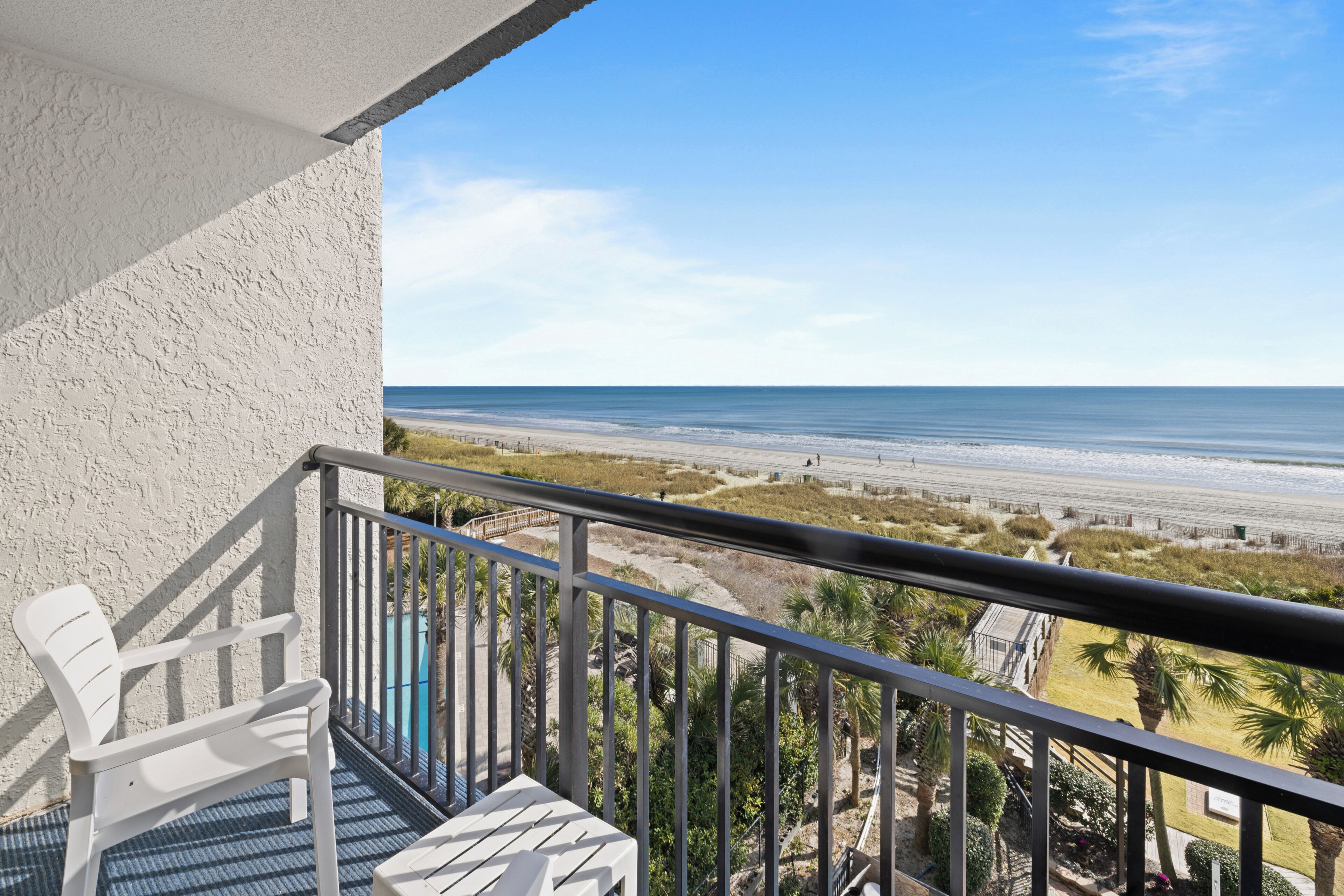 A balcony with a view of the beach, a white chair, and a table.