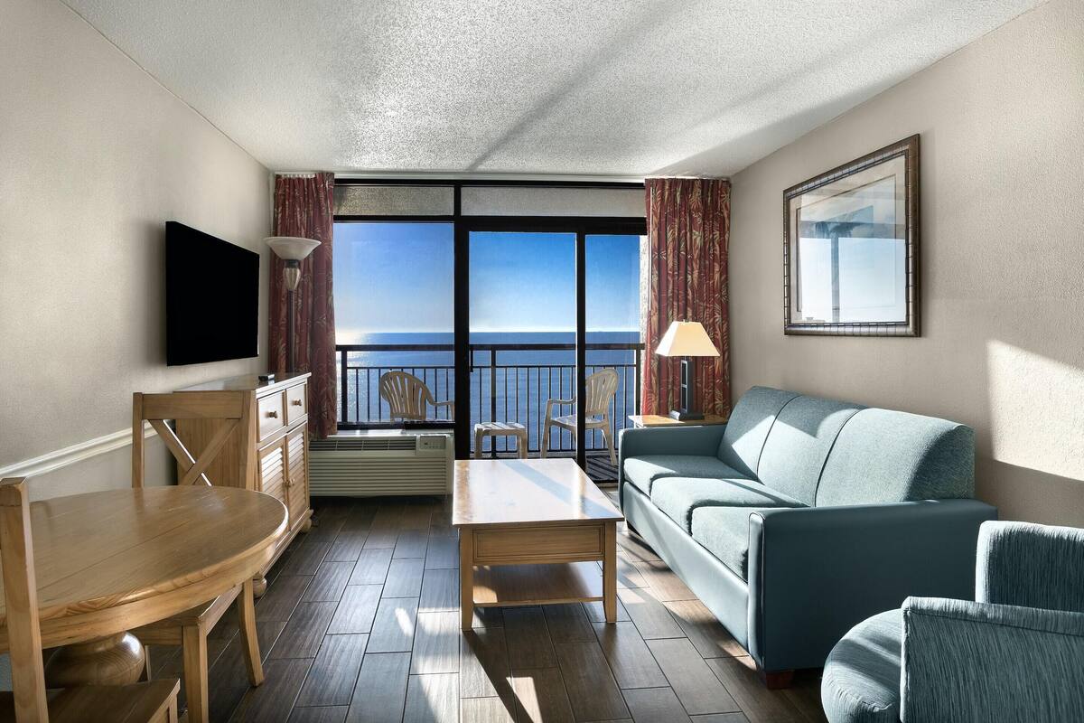 A modern hotel room with a balcony, a flat-screen TV, a wooden dining table, a sofa, and a lamp.