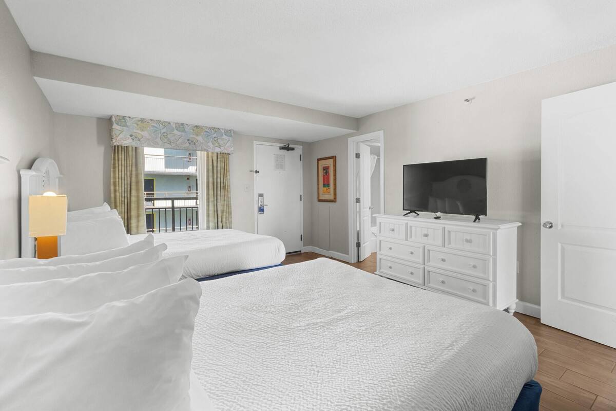 A hotel room with two beds, a TV, a dresser, and a balcony with curtains.