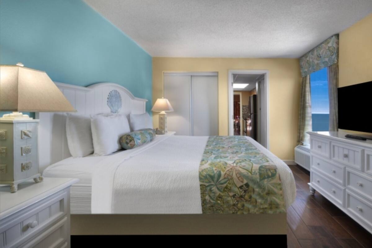 A hotel room with a bed, a television, a dresser, and a view of the ocean.