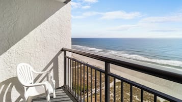 A balcony with a white plastic chair, overlooking a beach with waves and a clear sky.