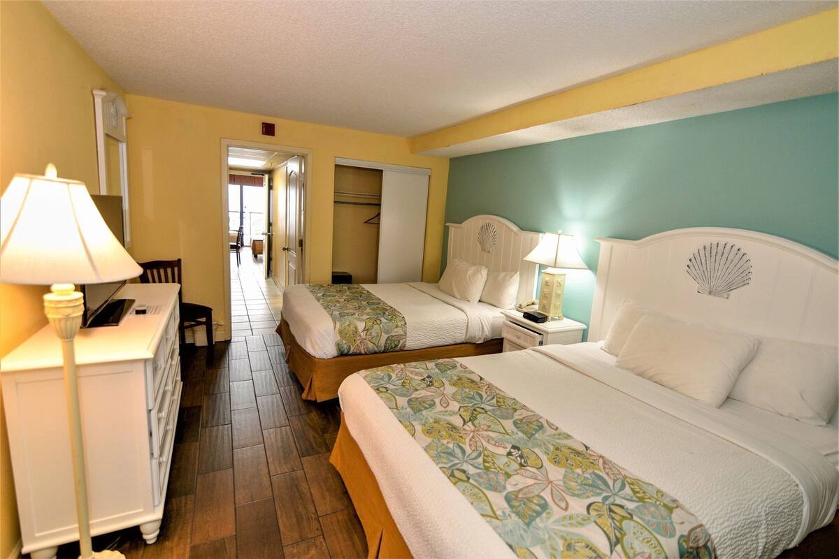 A hotel room with two beds, a desk, a chair, and a lamp.