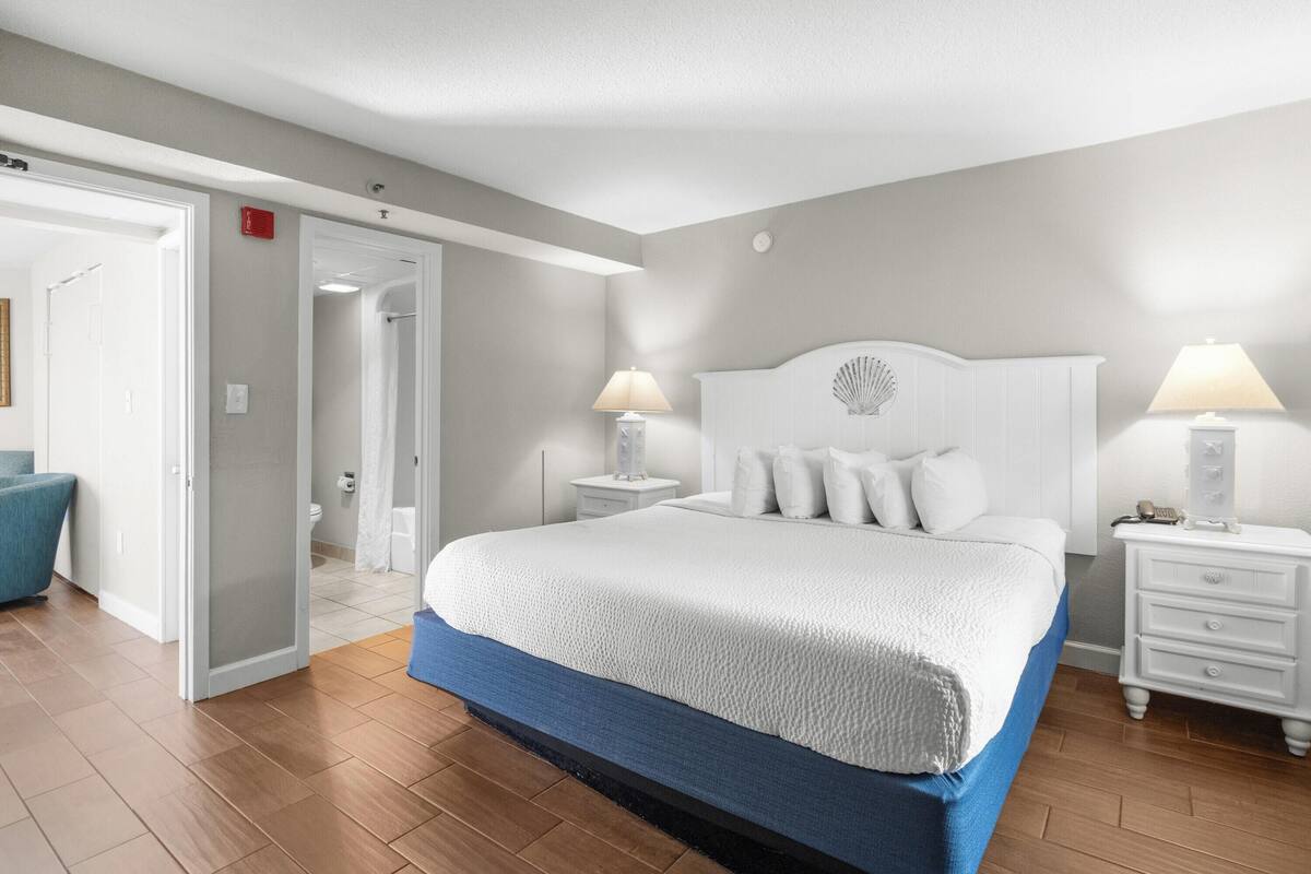 A bedroom with a large bed, two bedside tables with lamps, a white headboard, and a blue accent on the bed skirt.