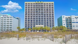 A high-rise hotel with multiple balconies, a beach in the foreground, and palm trees.