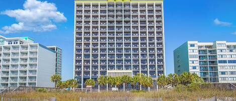 A high-rise hotel with multiple balconies, a beach in the foreground, and palm trees.
