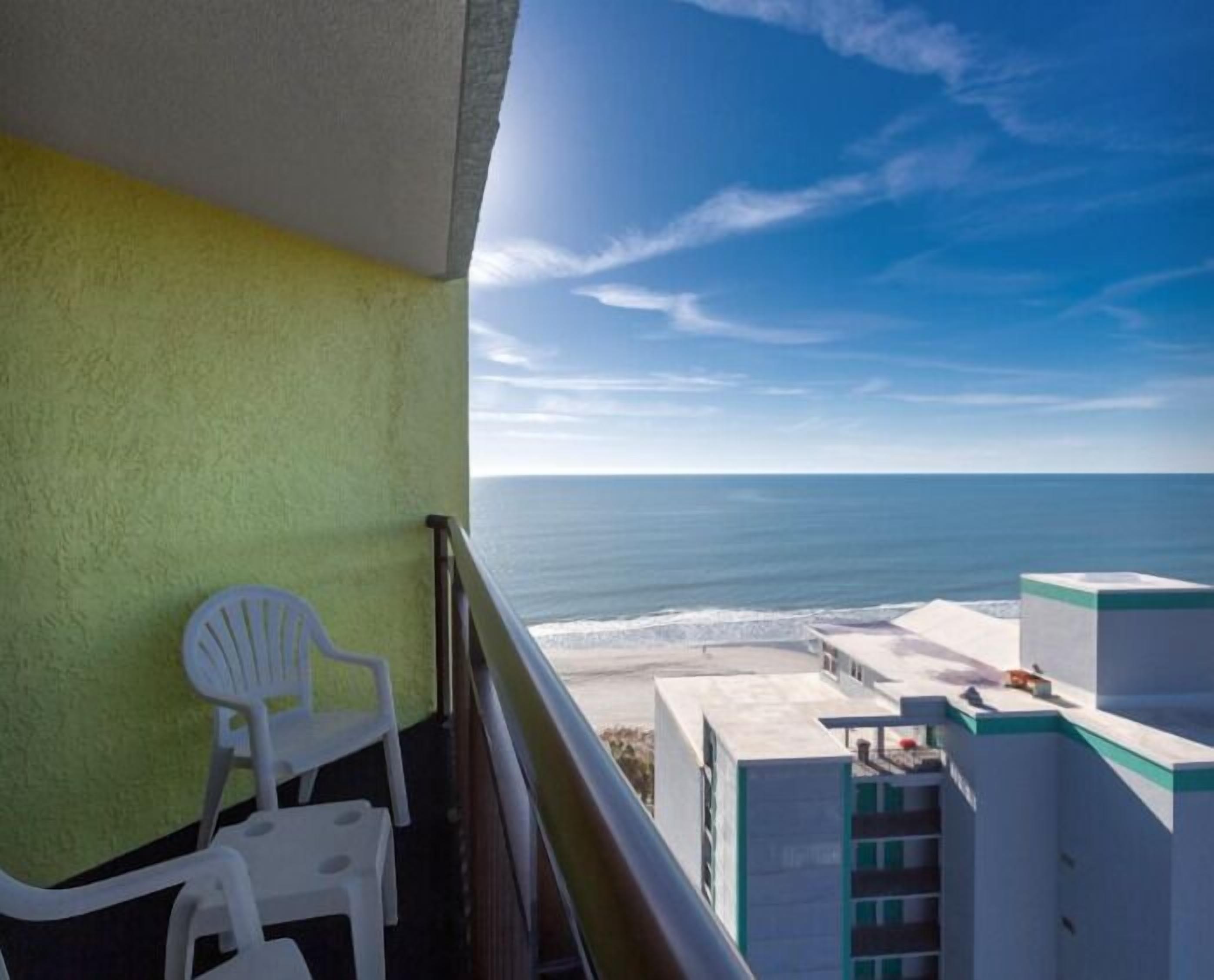 Un balcon donnant sur l’océan, une chaise blanche et un immeuble à la façade verte et blanche.
