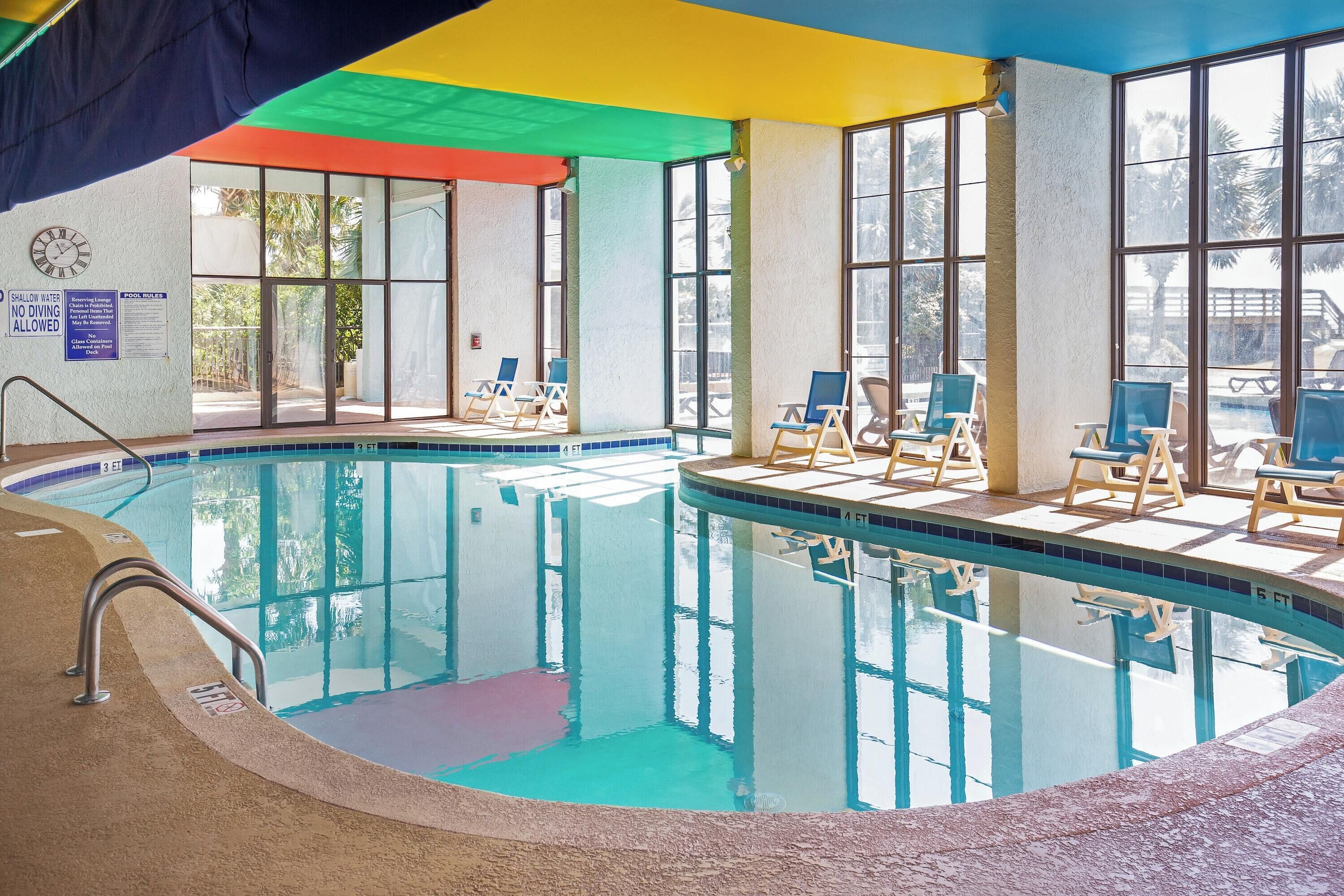 An indoor swimming pool with clear water, surrounded by lounge chairs and a covered area with a colorful roof.