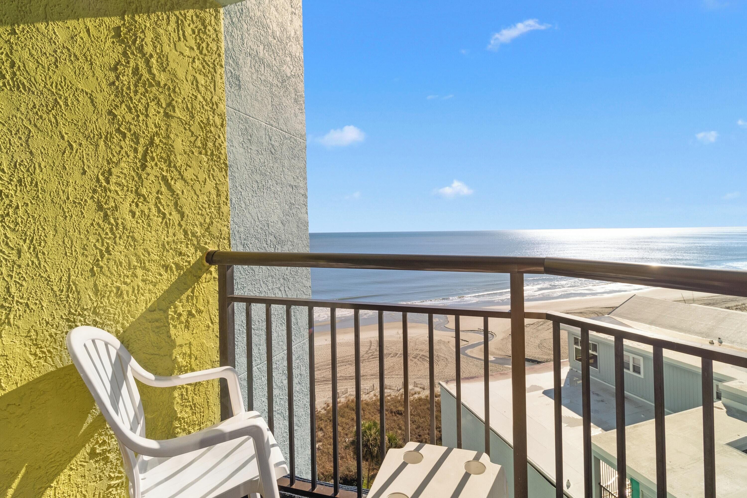 Un balcon avec vue sur l’océan, une chaise blanche et un bâtiment en arrière-plan.