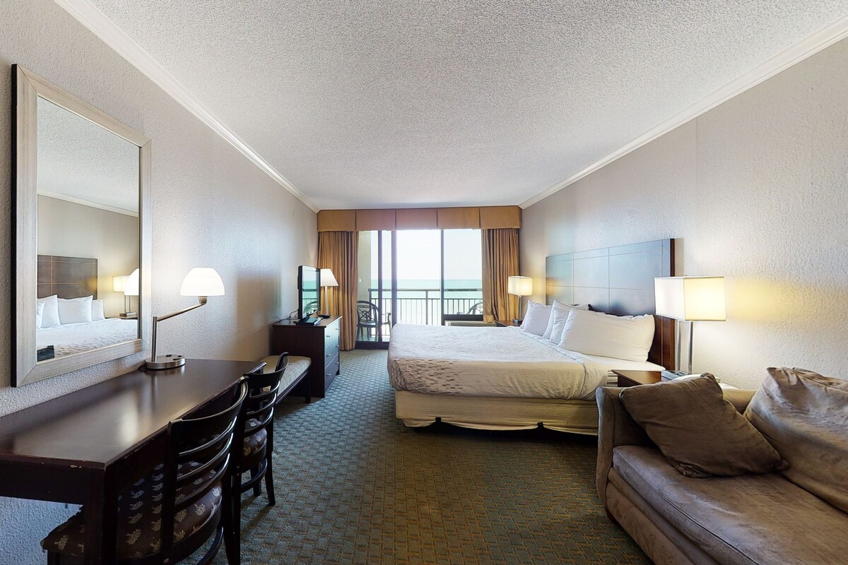 A hotel room with a bed, a desk, a chair, a sofa, and a large window offering a view.